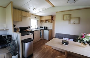 A compact kitchen with wooden cabinets, a microwave, and a sink. There is a dining table with a vase of flowers and a small trash bin. - Cozy Chalet With a View Over the Fields (Drachten)