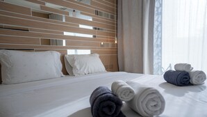 Una cama bien hecha con ropa de cama blanca y gris, cabecera de madera y toallas enrolladas sobre la cama.