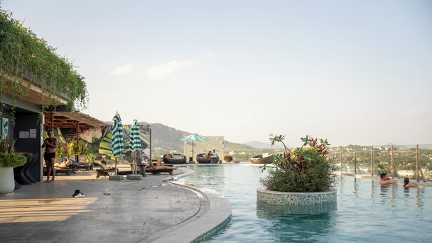 Una piscina infinita con sillones de descanso y sombrillas, rodeada de vegetación y con vistas a colinas distantes.