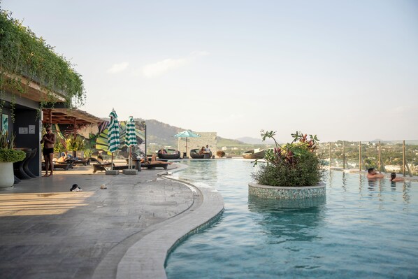 Ein Infinity-Pool mit Liegestühlen und Sonnenschirmen, umgeben von Grün und Blick auf ferne Hügel.