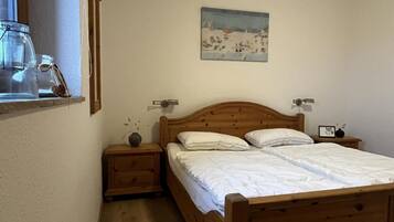 A bedroom with a wooden bed, a nightstand with a vase, a painting on the wall, and a window with a view of the outdoors.