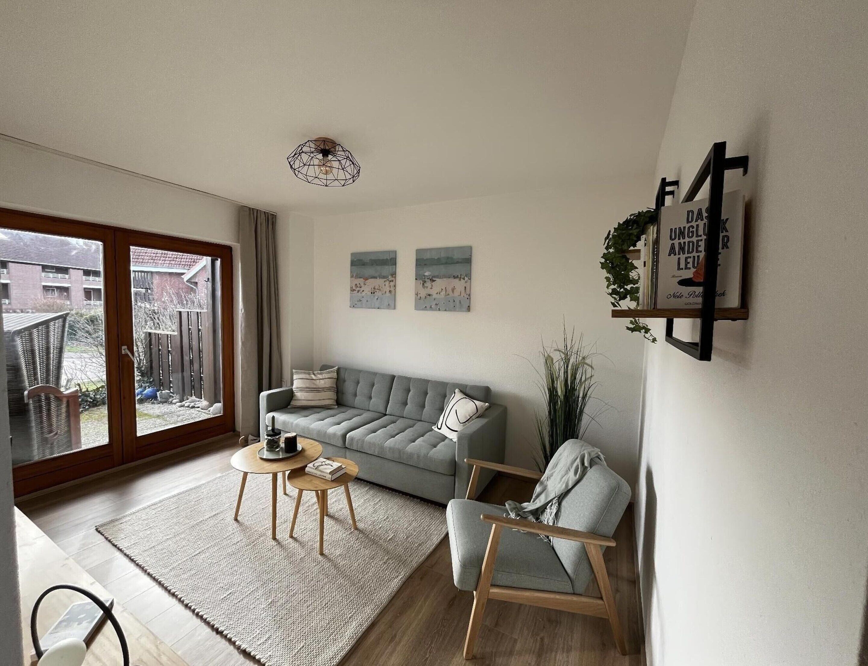 A modern living room with a grey sofa, a wooden chair, a small round table, and a large window with a view of a building and a fence.