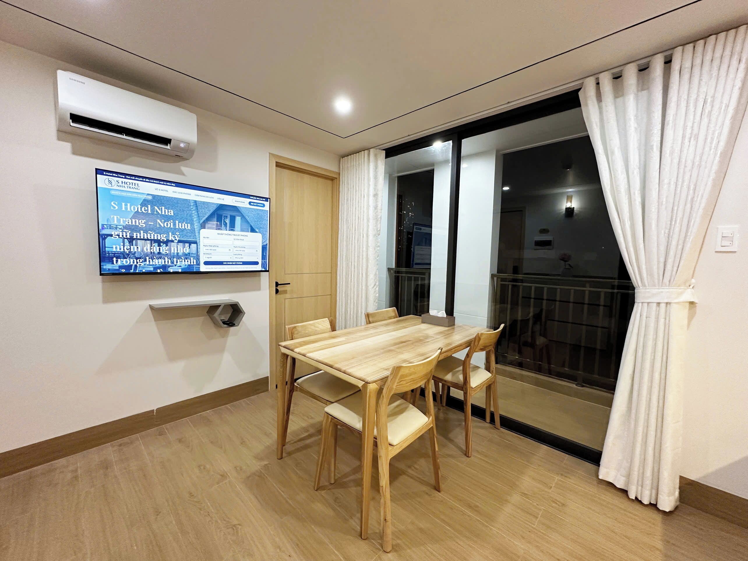 A hotel room with a wooden dining table, chairs, a TV mounted on the wall, and a sliding glass door leading to a balcony.