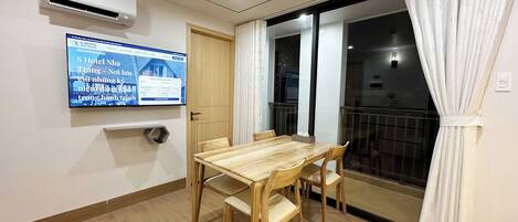 A hotel room with a wooden dining table, chairs, a TV mounted on the wall, and a sliding glass door leading to a balcony.