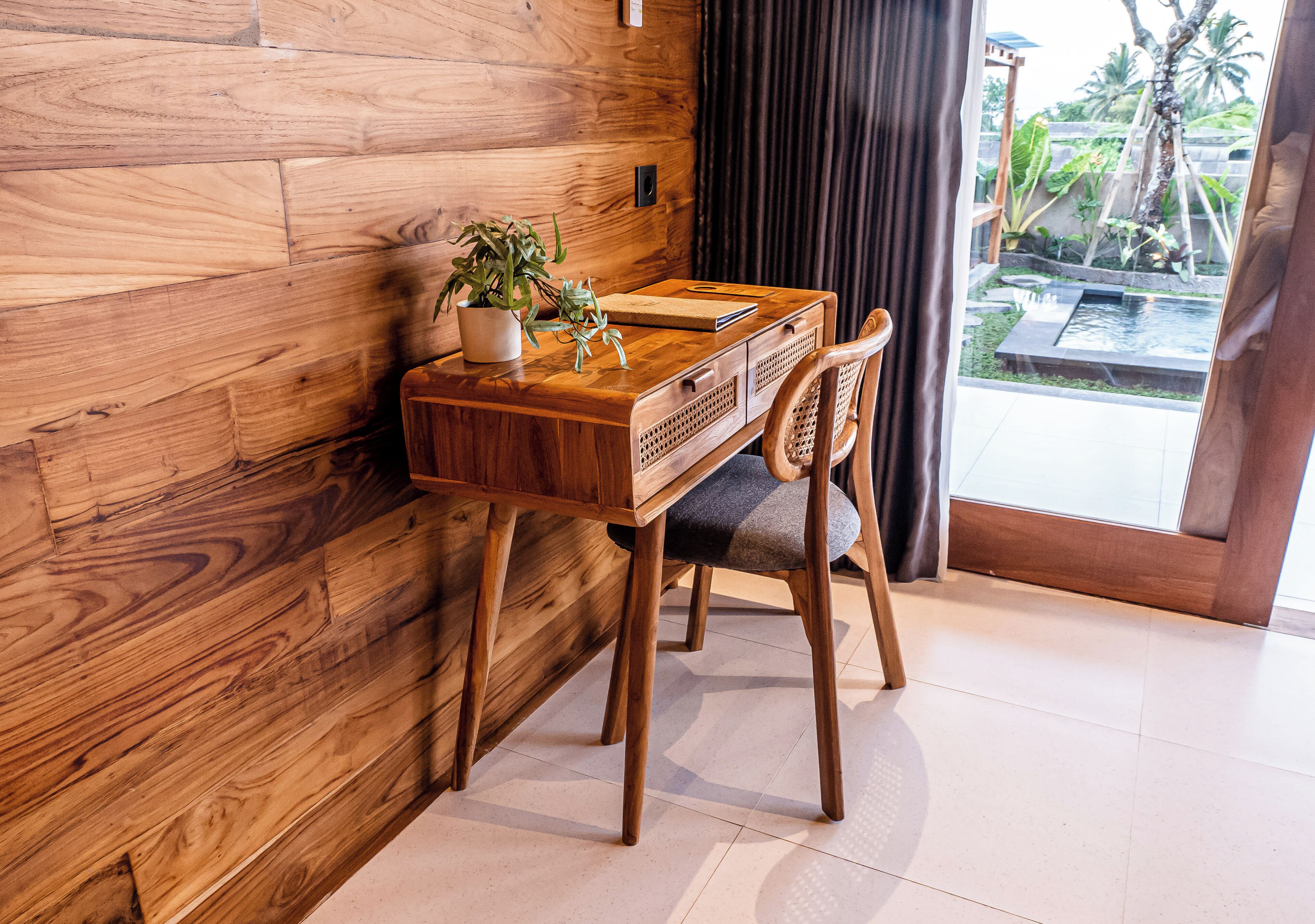 A wooden desk with a plant, a notebook, and a chair in a room with a wooden wall and a view of a garden through a glass door.