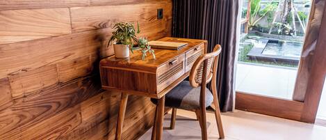 A wooden desk with a plant, a notebook, and a chair in a room with a wooden wall and a view of a garden through a glass door.