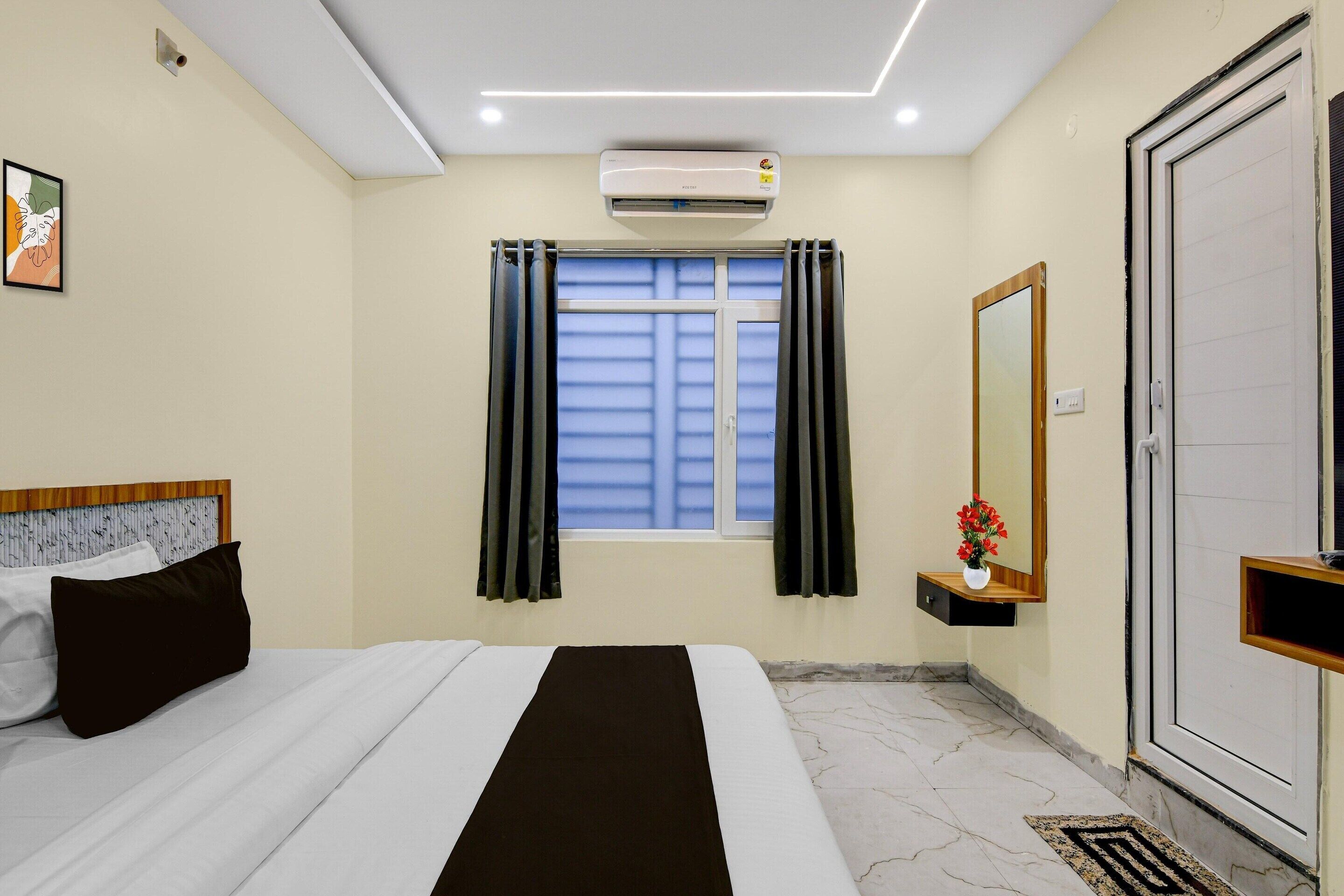 A hotel room with a bed, air conditioning unit, window with curtains, a bedside table with a vase, and a door.