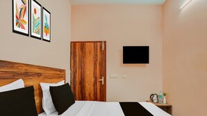 Una habitación de hotel con cabecera de madera, cama con ropa de cama blanca y negra, televisor de pantalla plana en la pared y una mesita con artículos de aseo.