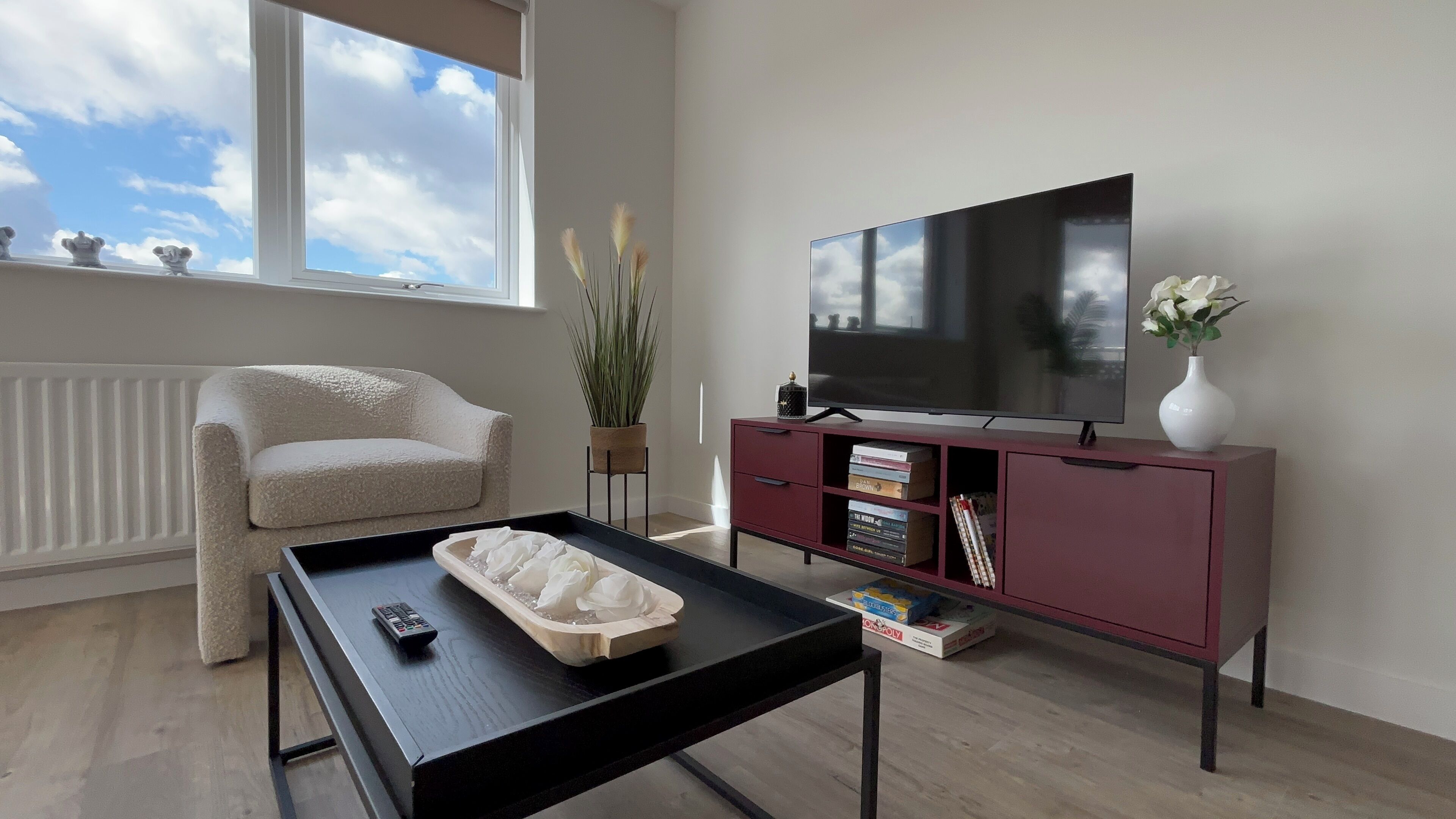 A living room with a sofa, a coffee table, a TV on a stand, a radiator, and a window with a view of the sky.