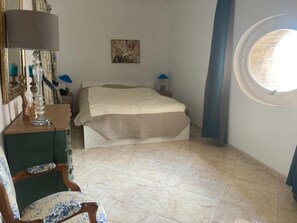 Ein Schlafzimmer mit einem Bett, einem Sessel, einer Kommode, einer Lampe, einem Gemälde und einem Fenster.