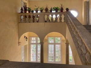 Ein mehrgeschossiges Interieur mit einer Steintreppe, Bogenportalen und einem Balkon mit Topfpflanzen.