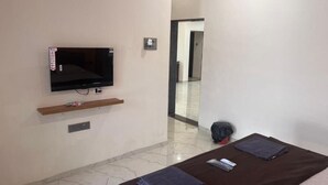 Hotellihuone, jossa on sänky, seinälle asennettu televisio, pieni hylly ja ovi, joka johtaa toiseen huoneeseen.