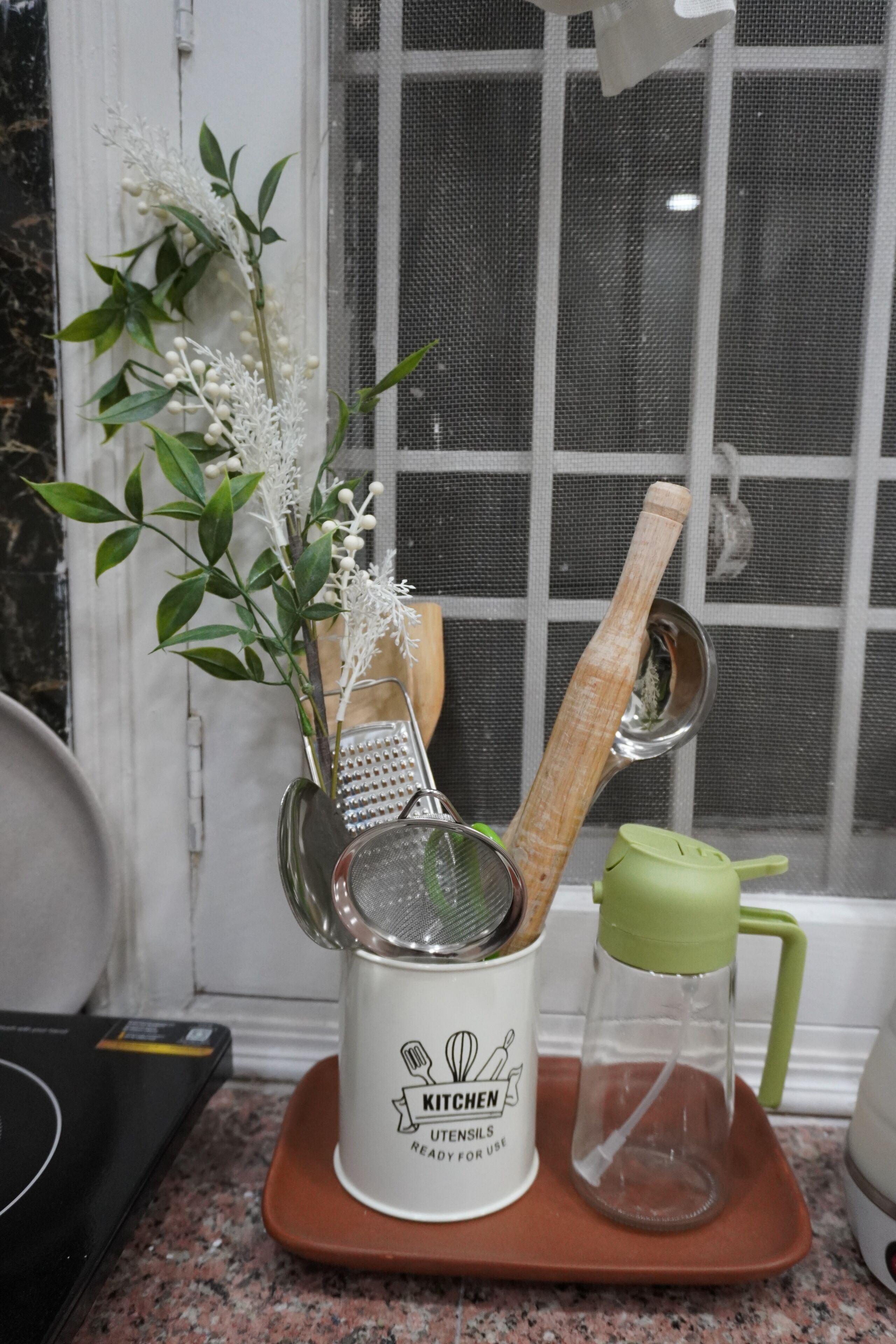 A kitchen counter with a white mug labeled 'KITCHEN UTENSILS READ FOR USE,' a green water bottle, a wooden spoon, a metal strainer, and a plant with green leaves and white berries.