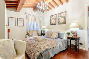 Ein Schlafzimmer mit Bett, Stuhl, einer Kandelaber und gerahmten Kunstwerken an der Wand.