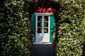Ein Weg führt zu einem Haus mit weißer Tür und grünen Fensterläden, umgeben von üppigem Grün.