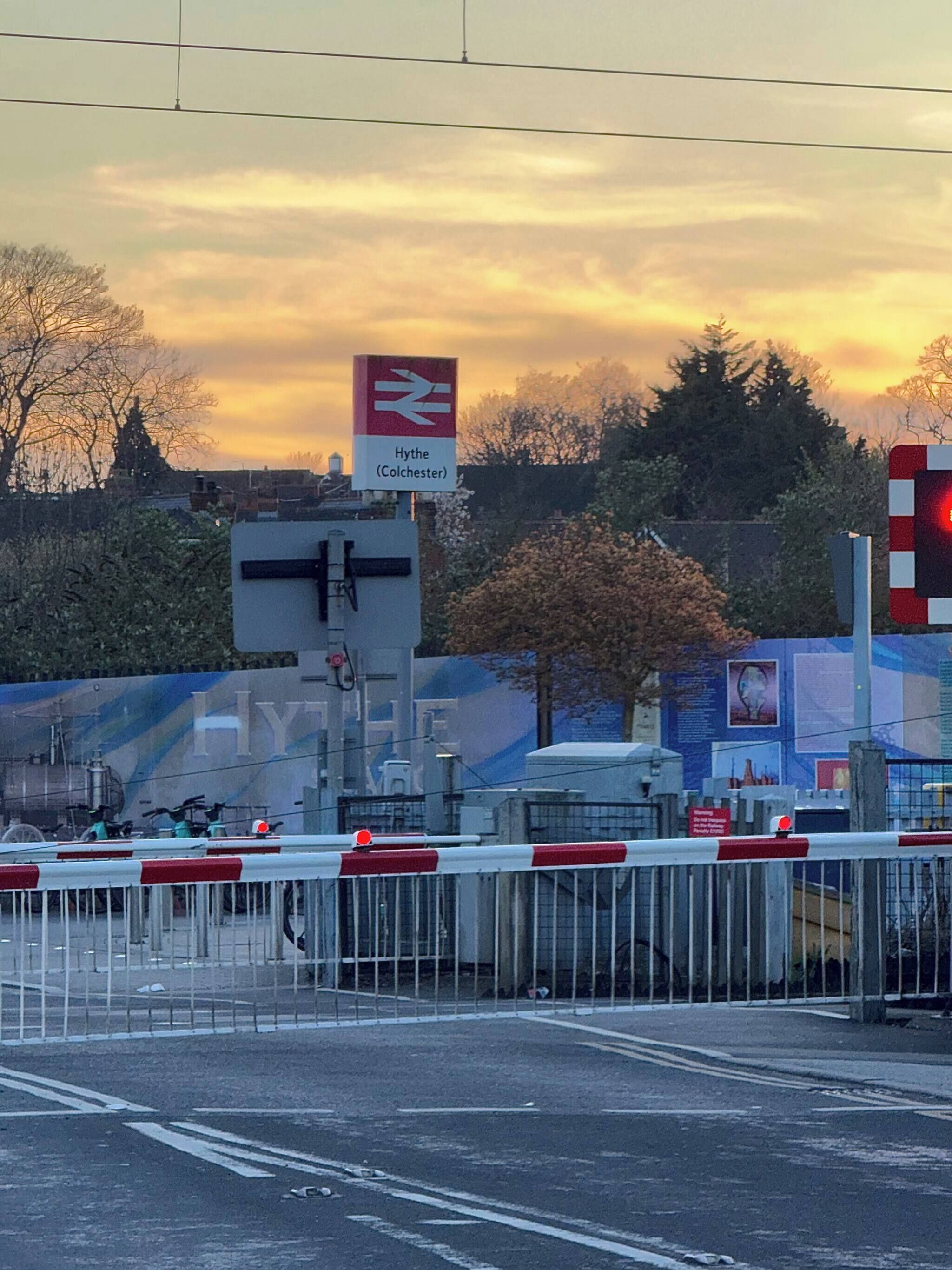 A railway station platform with a sign for Hythe (Colchester), a barrier gate, and a red signal light.