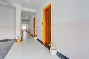 Un pasillo largo y estrecho con paredes blancas y piso de mármol, que conduce a varias puertas con marcos de madera y detalles en naranja.