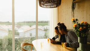 A dining area with a large window, a wooden table, and two people seated. - 4-bedroom Stylish Bungalow by the Sea ('s-Gravenzande)