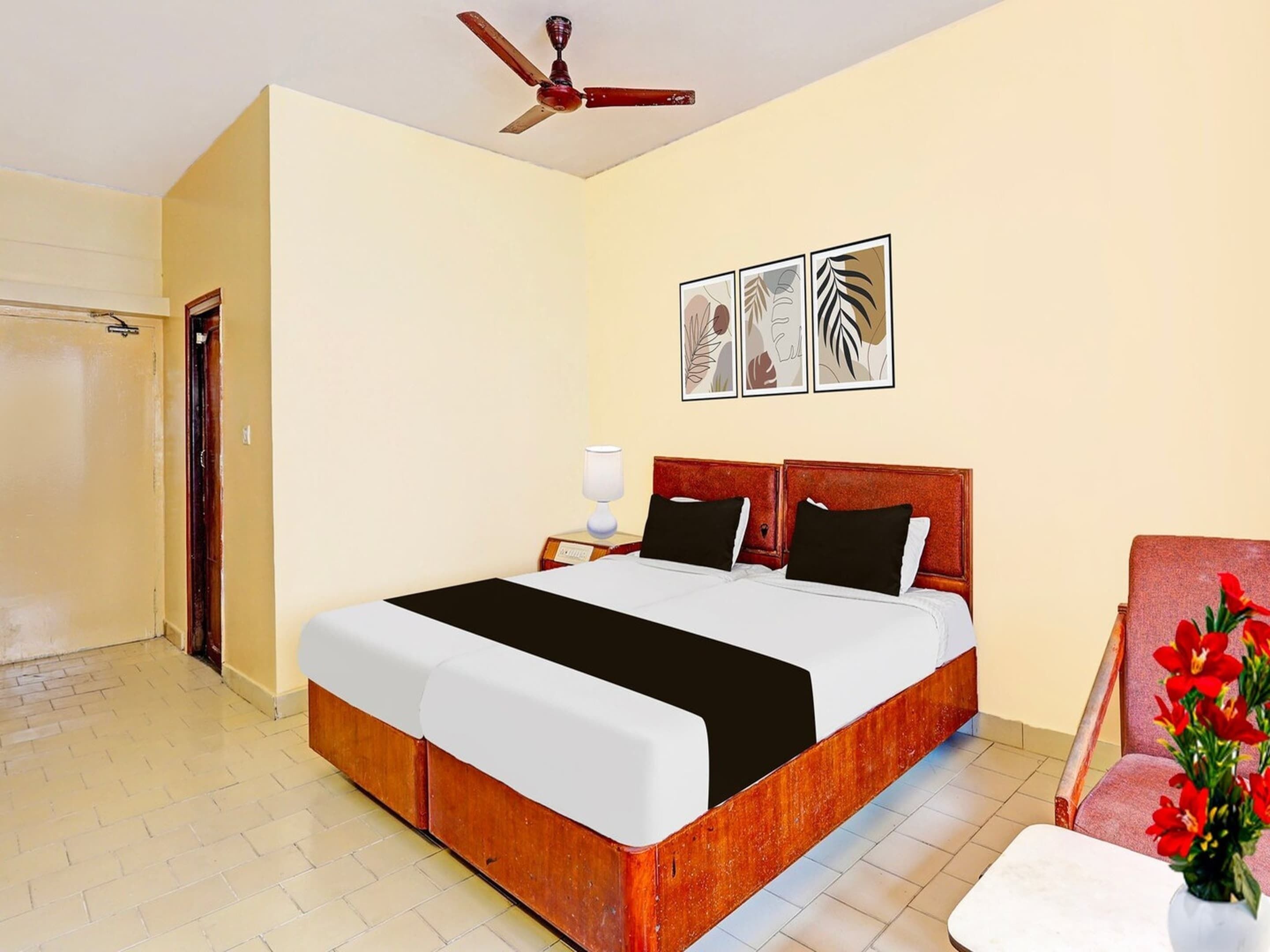 Una habitación de hotel con una cama, una mesita de noche, un ventilador de techo, una puerta y una pared decorada con cuadros.