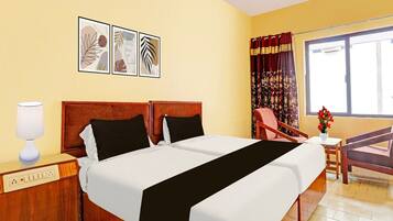 Una habitación de hotel con una cama doble, una mesita de noche con una lámpara, una silla, una pequeña mesa con un jarrón y una ventana con cortinas.