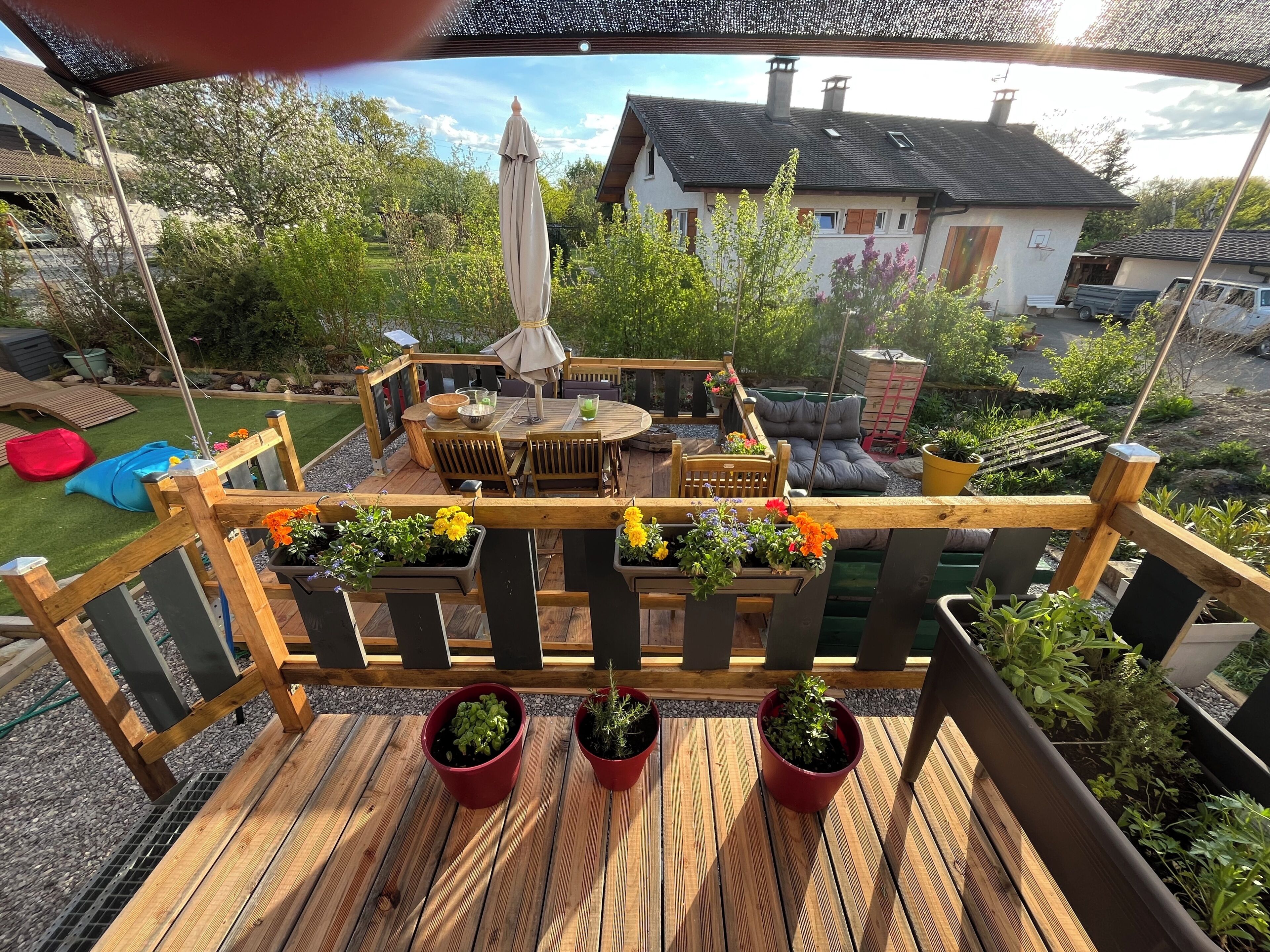 Une terrasse en bois, agrémentée de meubles d’extérieur, de plantes en pot et offrant une vue sur une maison en arrière-plan.