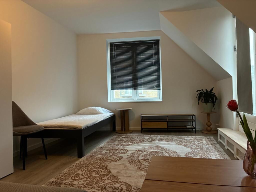 Ein kleiner, gut beleuchteter Raum mit einem Bett, einer Bank, einem Tisch, einer Topfpflanze und einem Fenster mit Jalousien.
