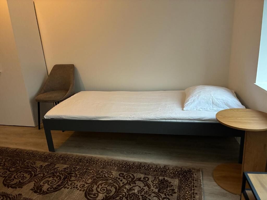 A single bed with a white mattress and a dark frame, a small round wooden table, a patterned rug, and a chair.