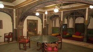 A room with a round table, wooden chairs, a sofa with cushions, and ornate wall decorations. - Experience Royal Living in a Heritage Haveli (Jaipur)