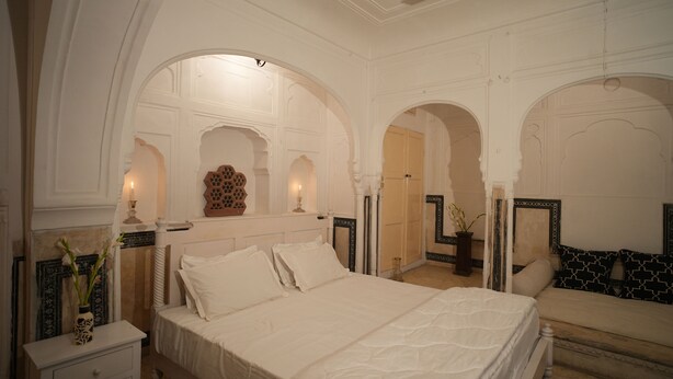 A bedroom with a large bed, a nightstand, a bedside table, and a mirror. - Experience Royal Living in a Heritage Haveli (Jaipur)