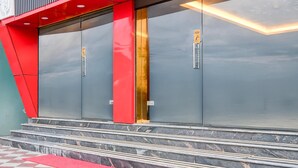 A modern building with a red facade, large glass doors, and a set of stairs leading to a red carpet.