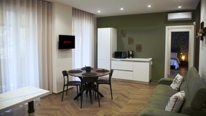 Ein modernes Wohnzimmer mit Esstisch, Stühlen, einer Couch, einem Fernseher, einer Küchenzeile und einem durchgehenden Gang sichtbaren Schlafzimmer.