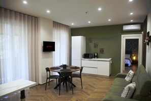 Ein modernes Wohnzimmer mit Esstisch, Stühlen, einer Couch, einem Fernseher, einer Küchenzeile und einem durchgehenden Gang sichtbaren Schlafzimmer.