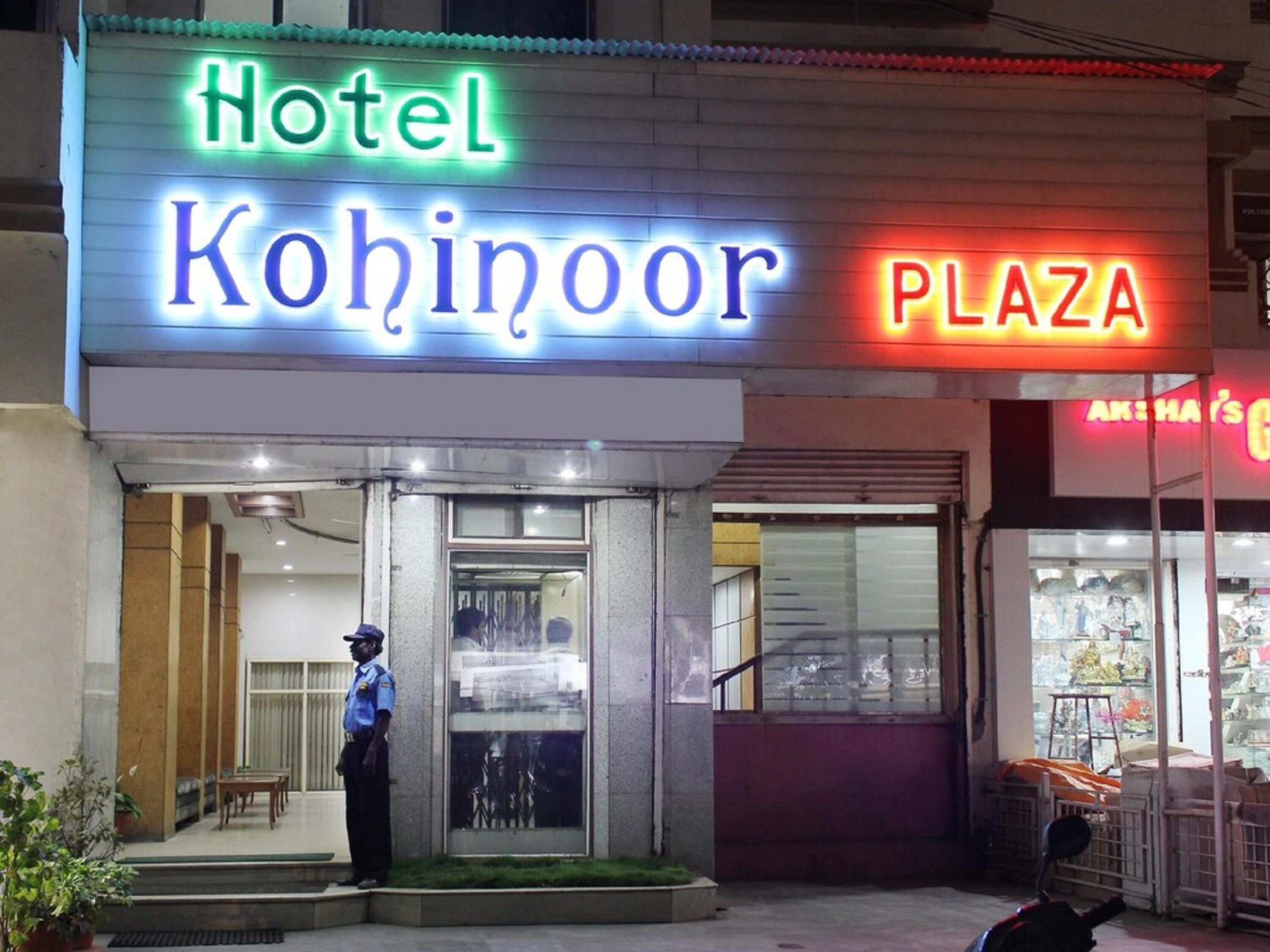 A hotel named 'Hotel Kohinoor' with a neon sign, a security guard, and a storefront labeled 'PLAZA' in the background.