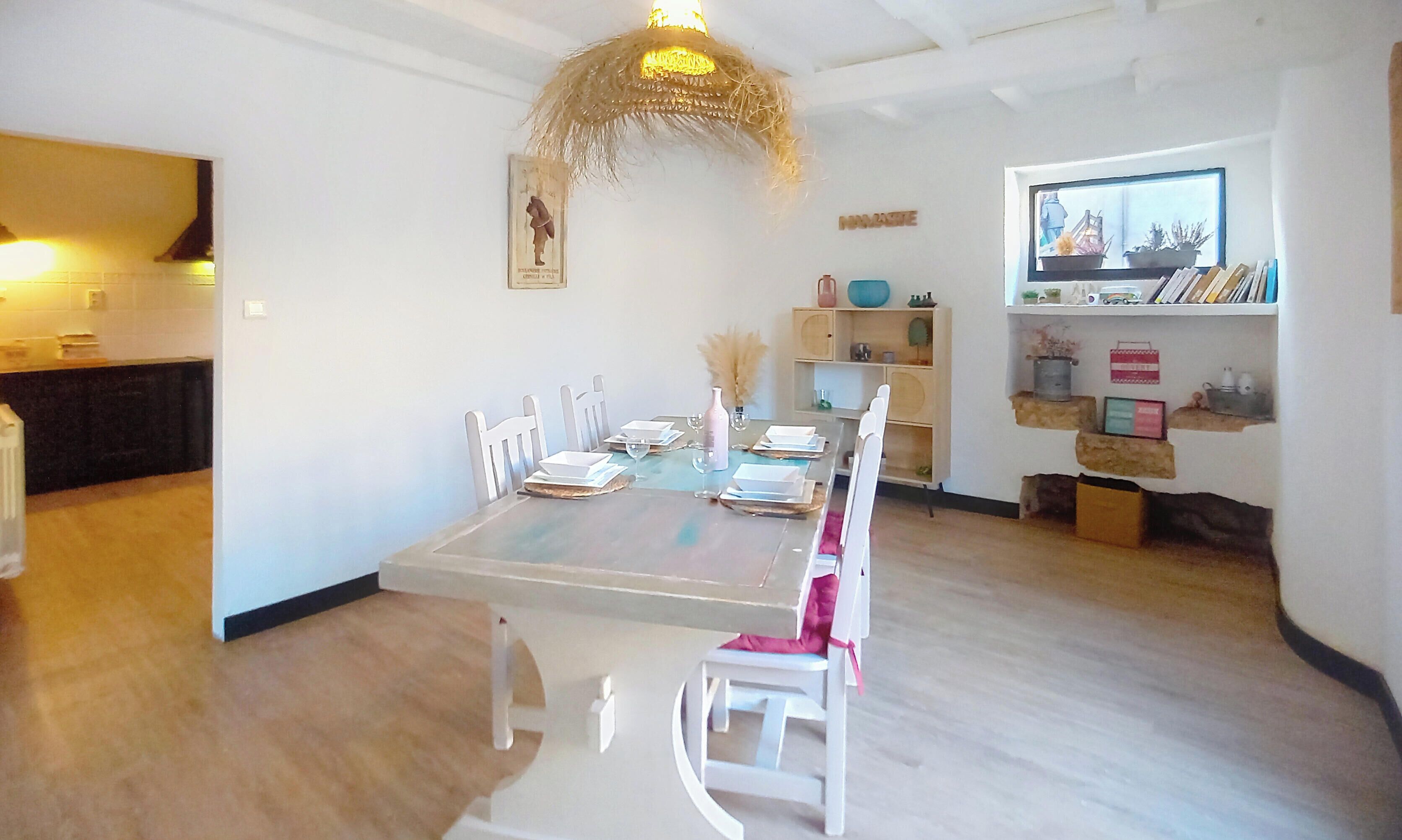 A dining area with a long table, white chairs, a chandelier, and a kitchen in the background.
