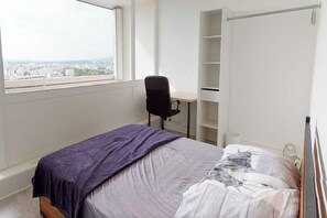 A bedroom with a bed, a desk, and a view of a cityscape through a window. - Private Room in a shared flat – Villeurbanne (Villeurbanne)
