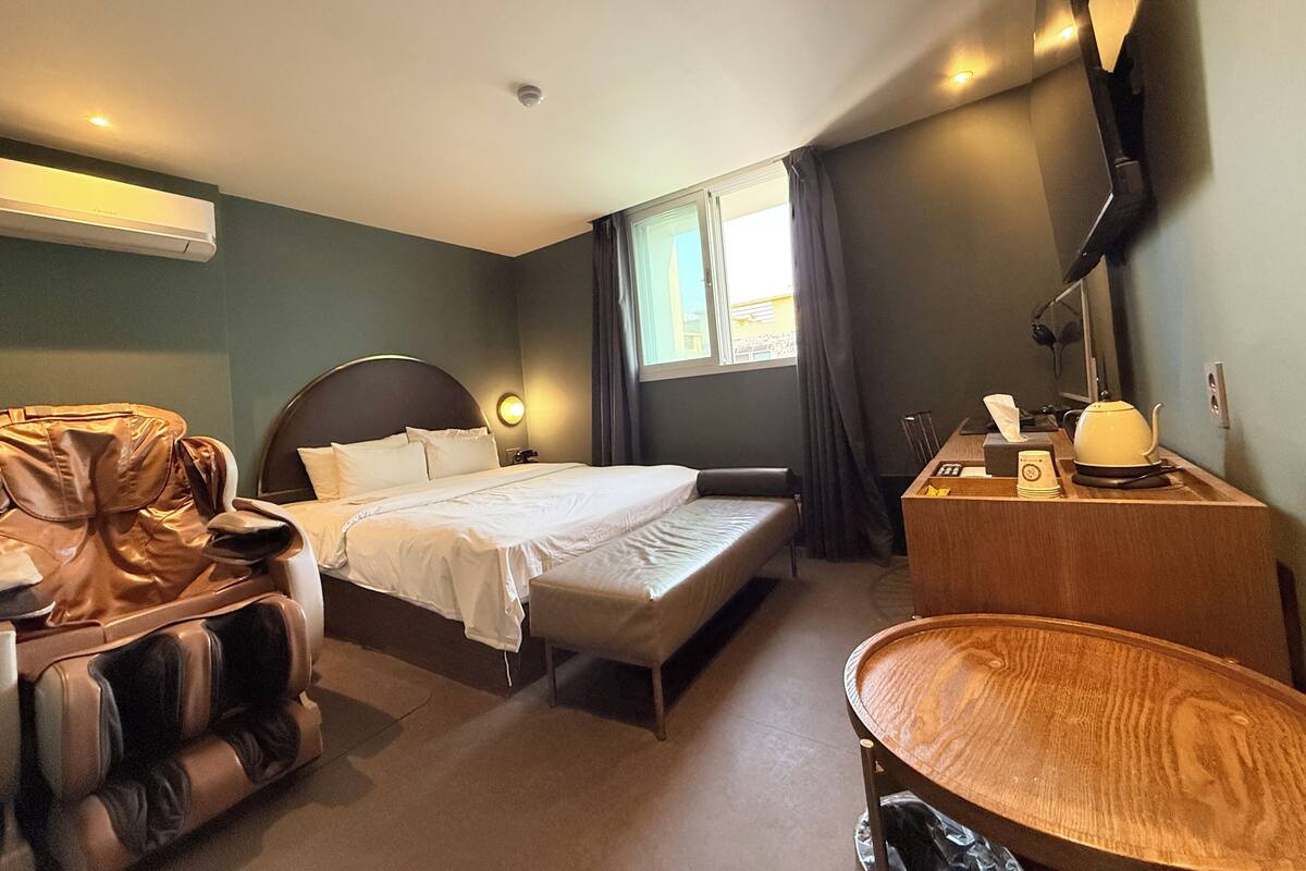 A hotel room with a bed, a massage chair, a desk with a lamp, and a TV on the wall.