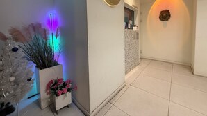 A hotel reception area with a sign reading 'HOTEL FLEX9', a reception desk, a decorative wall with a wreath, and a plant with pink flowers. - Flex9 (Uijeongbu)