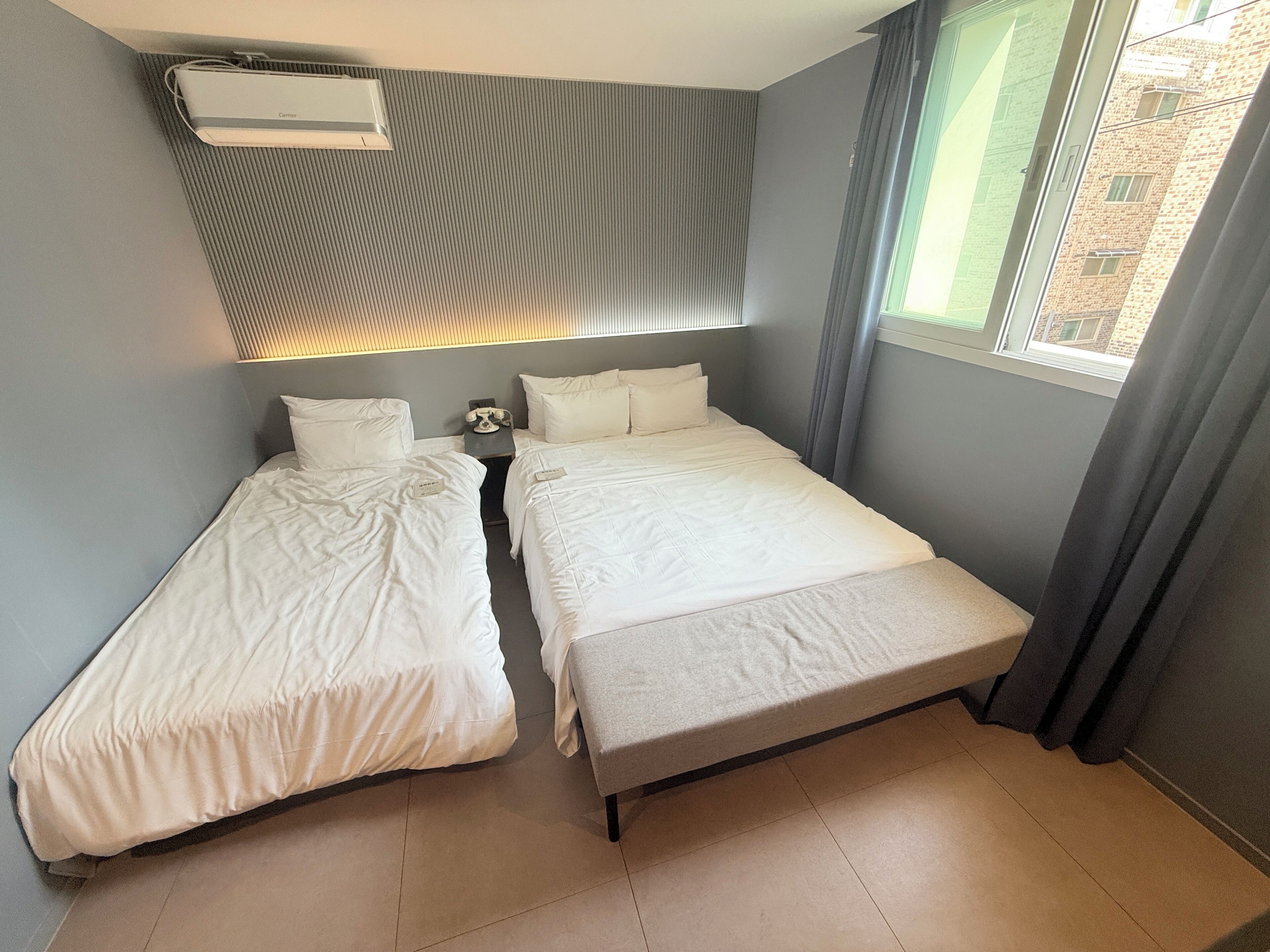 Quarto duplo com duas camas, mesa de cabeceira, ar condicionado e janela com cortinas.