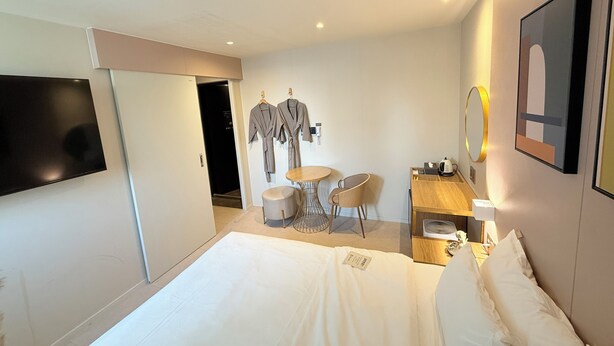 A hotel room with a bed, a TV, a desk with a chair, a small table, and a mirror. - Flex9 (Uijeongbu)