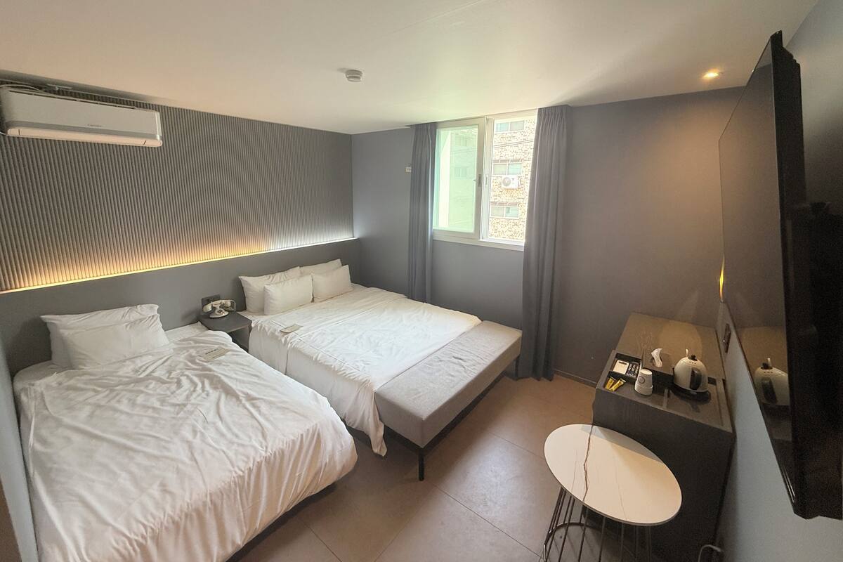 A modern hotel room with two beds, a small desk with a chair, a window with curtains, and a mounted television.