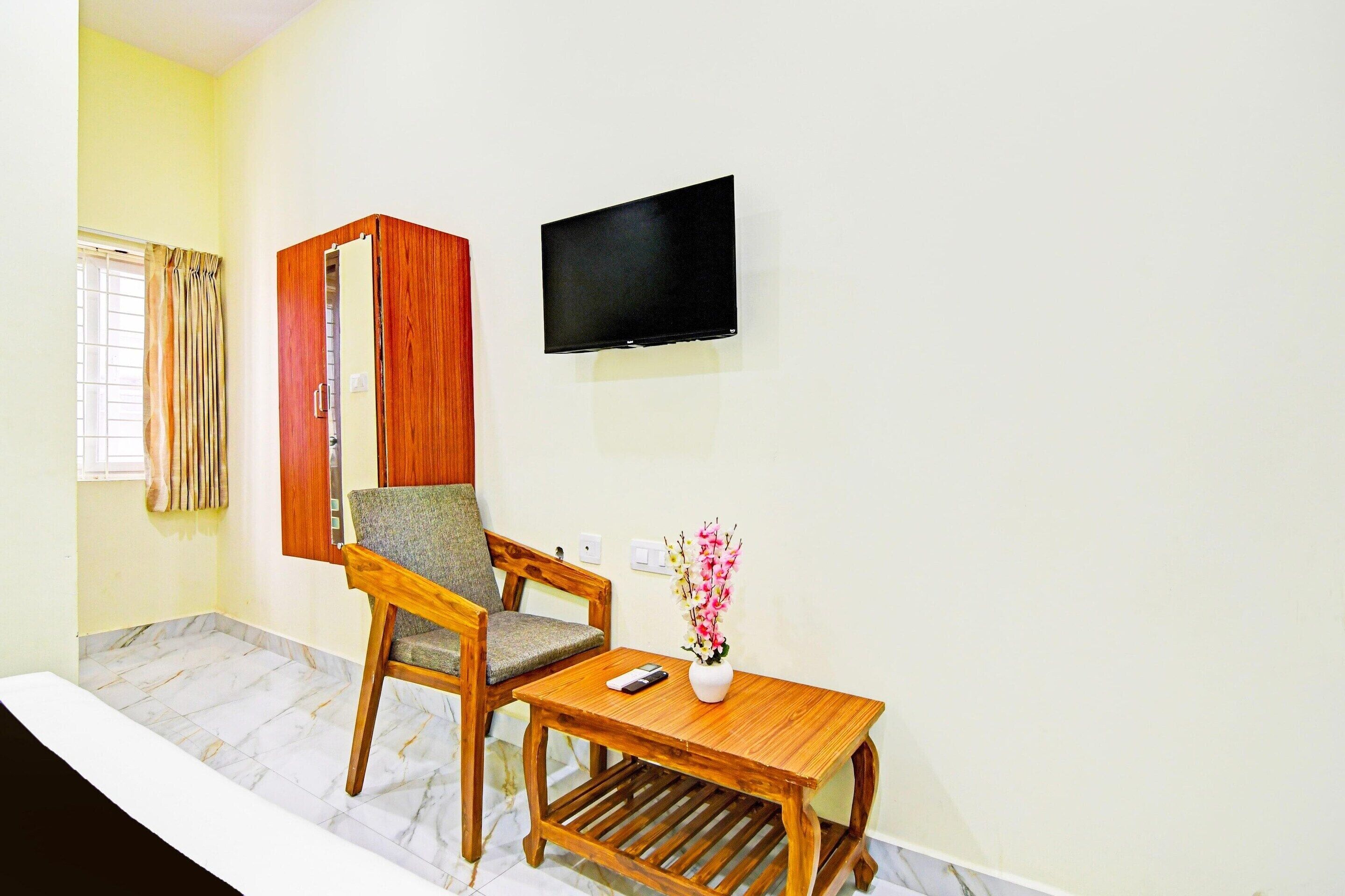 Una habitación con una silla de madera, una pequeña mesa de madera, un televisor de pantalla plana montado en la pared y una ventana con cortinas.