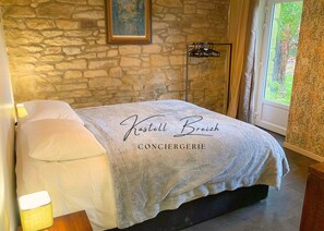 A bedroom with a stone wall, a bed with a grey blanket, a painting, and a window with curtains.