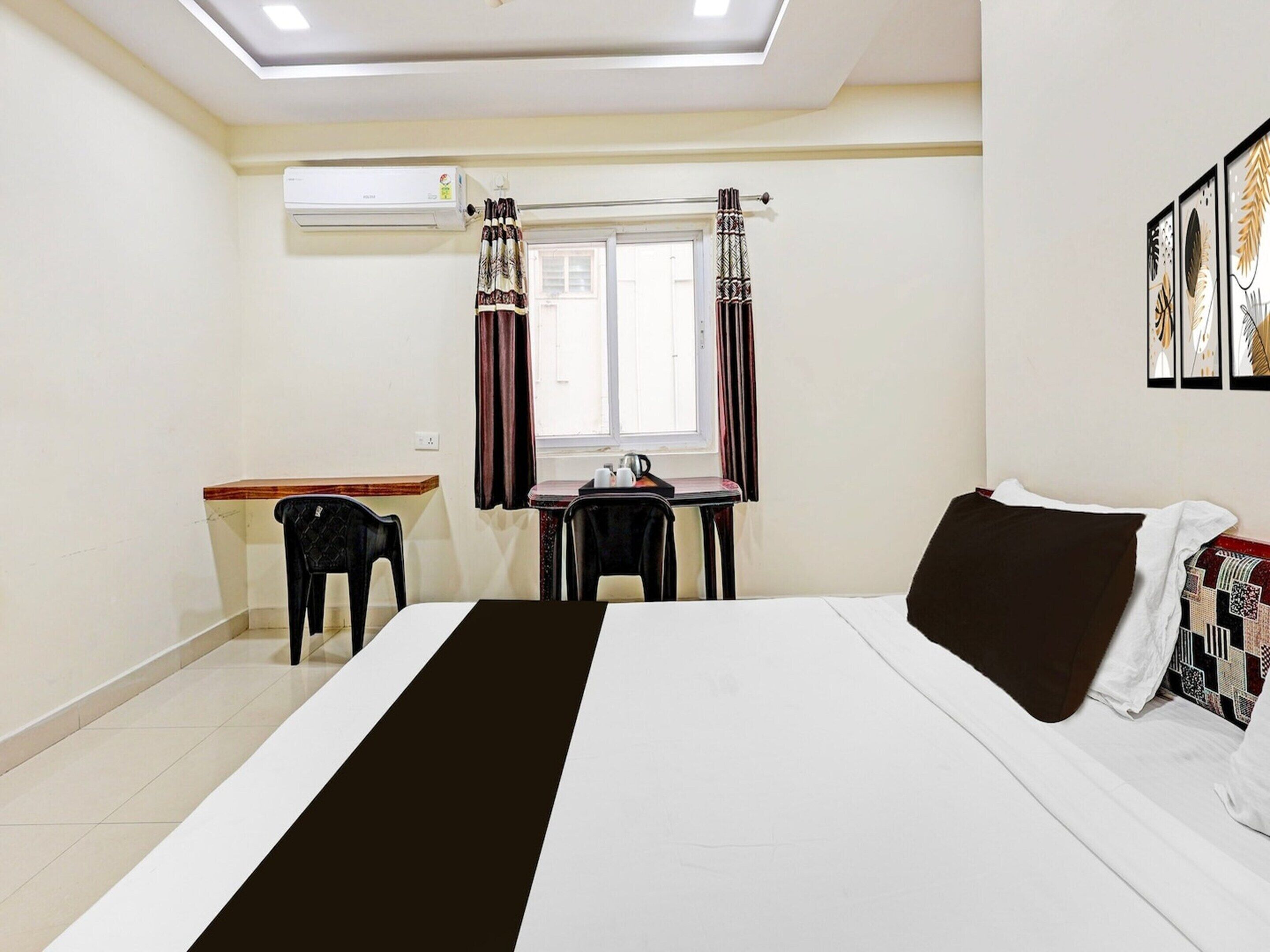 A hotel room with a bed, a desk, two chairs, a window with curtains, an air conditioning unit, and wall art.
