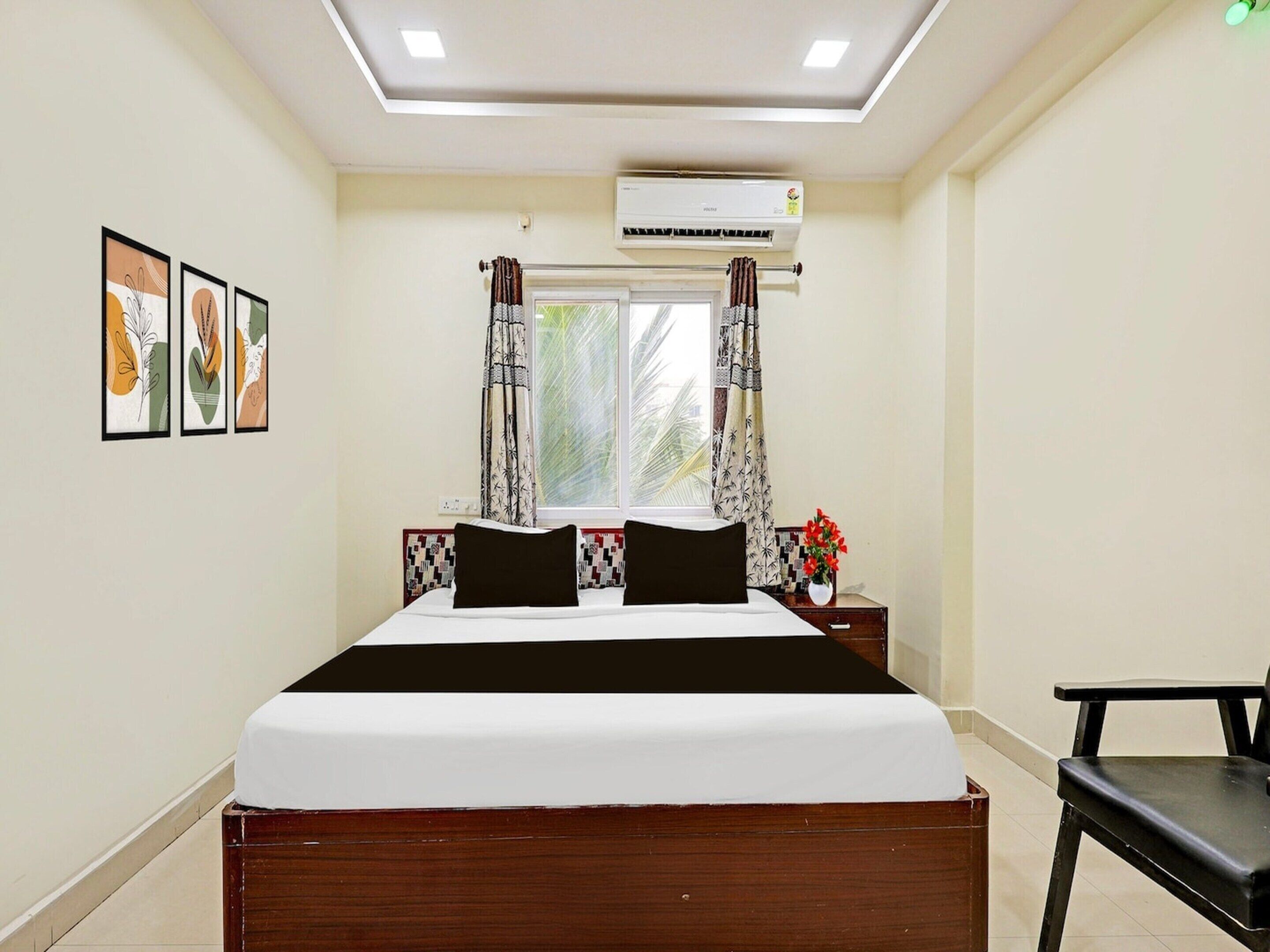 A bedroom with a bed, an air conditioner, a window with curtains, a nightstand, a chair, and framed artwork on the wall.
