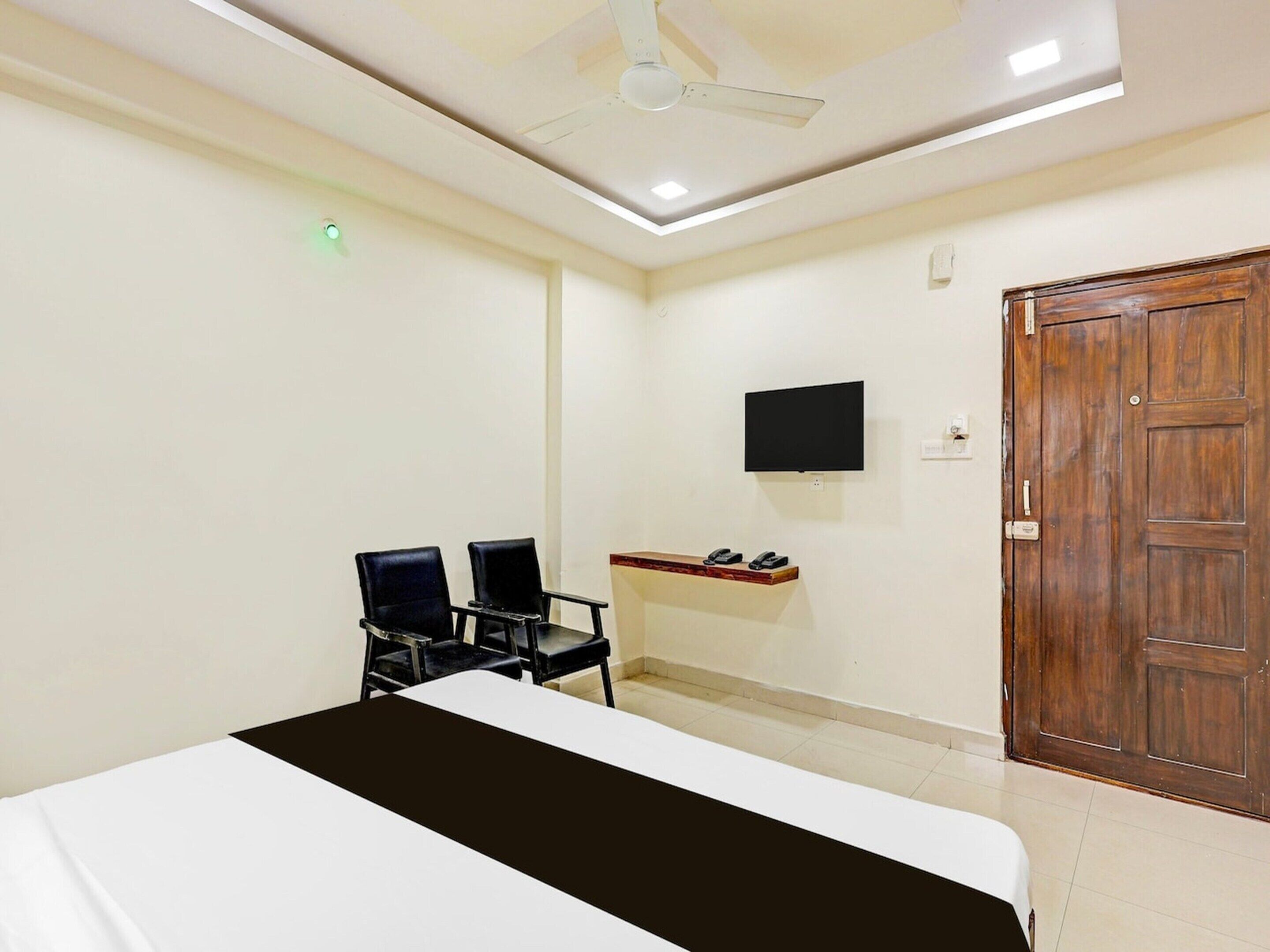 A room with a bed, two chairs, a TV, a wooden door, and a ceiling fan.