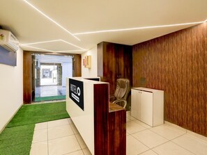 A hotel reception area with a reception desk, a leather chair, a small cabinet, and a wooden wall. - Premiumnana Chiloda Near Airport (Gandhinagar)