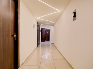 A hallway with a wooden door on the left, a wall-mounted light fixture, and a door at the end. - Premiumnana Chiloda Near Airport (Gandhinagar)