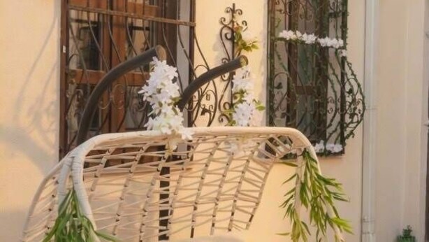Una silla colgante de mimbre con cojines, una ventana con rejas y una lámpara fijada a la pared.