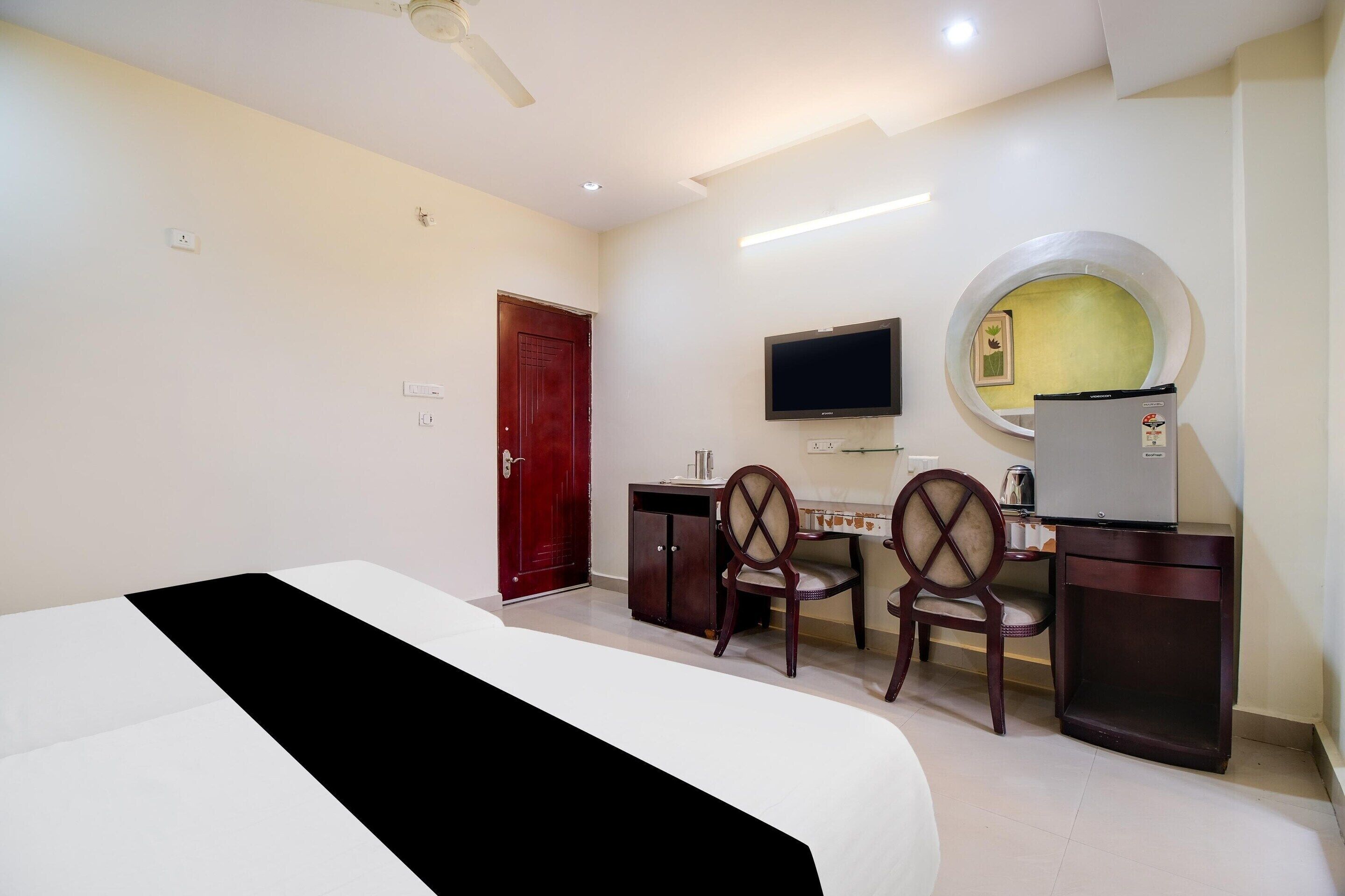 A hotel room with a bed, desk, chairs, a television, a refrigerator, and a door.