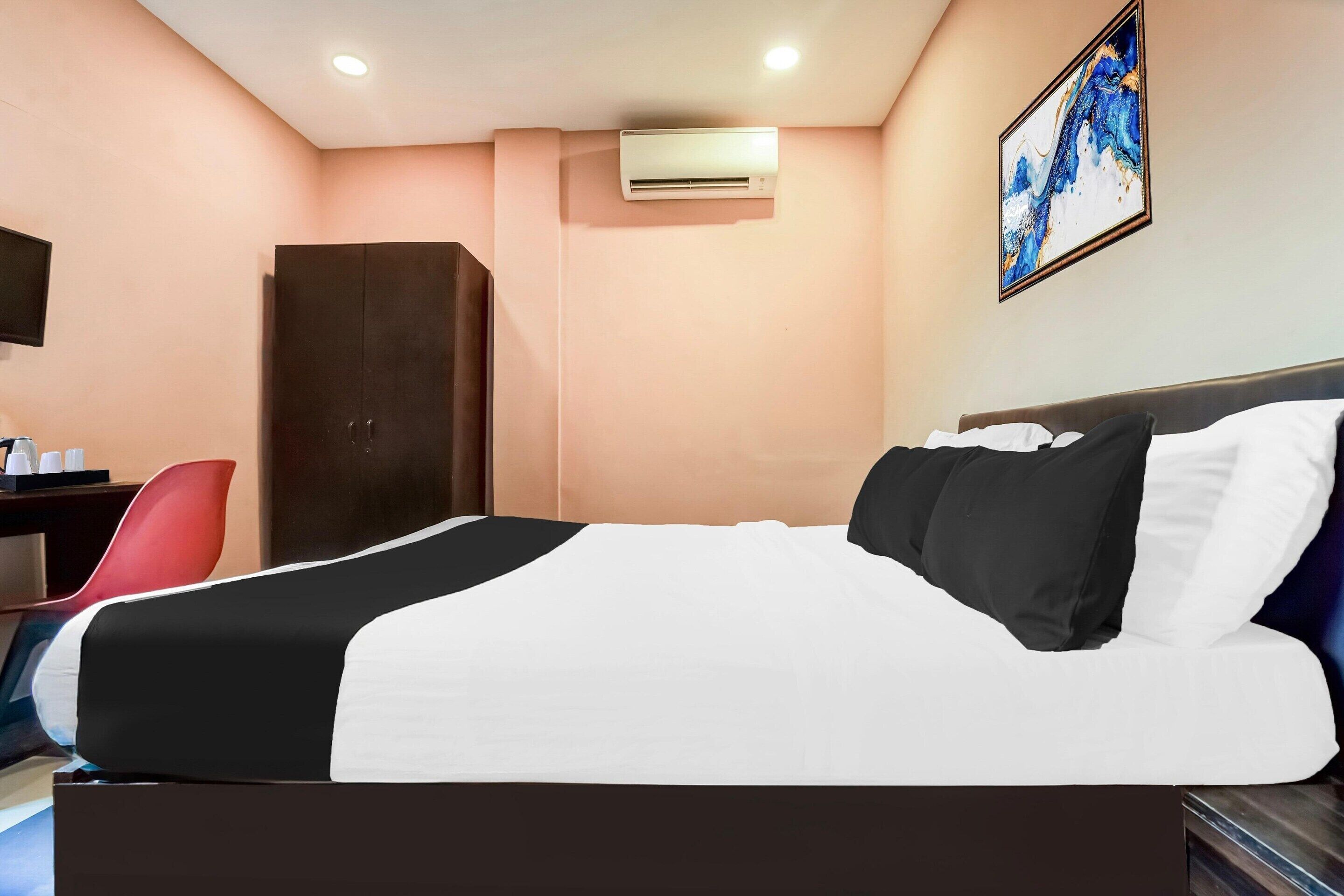 A hotel room with a bed, a desk, a chair, an air conditioning unit, and a painting on the wall.